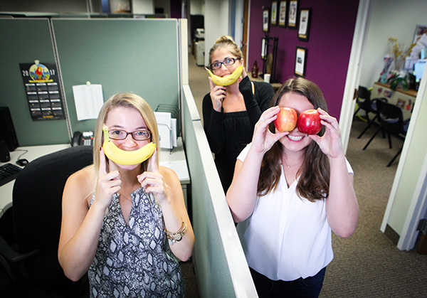 Happy employees holding bananas up to their faces as smiles and apples as eyes in the office