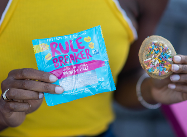 Woman holding cookie and packaging, which reads "Rule Breaker: Gluten-free & Vegan, Birthday Cake, Free From Top 8 Allergens, No Nuts!, Great for School!"