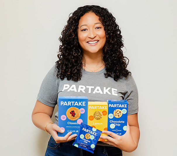 Smiling woman in Partake shirt holding boxes of Partake cookies