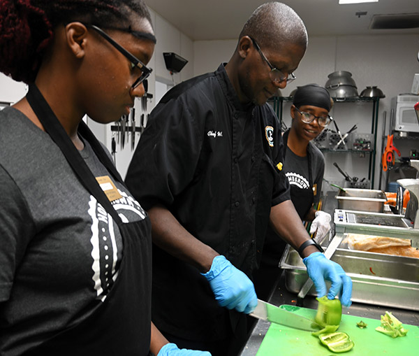 Man demonstrating chopping peppers in a commercial kitchen for two onlookers