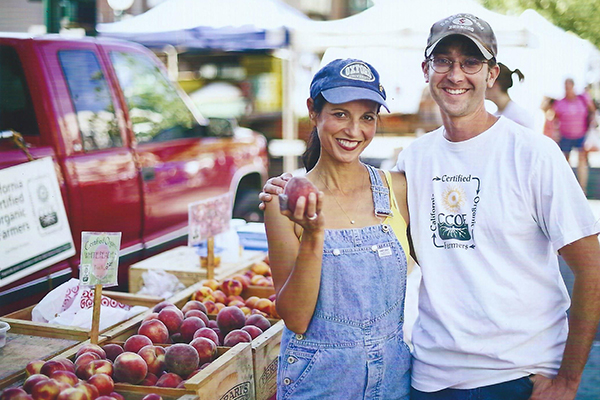 Smiling man and woman holding a peach at a farmers market