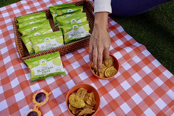 Hand reaching into a bowl of plantain strips, more Artisan Tropic plantain strips in bags in the background