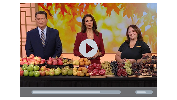 Two news anchors and a woman representing The FruitGuys standing in front of a table of fresh fruit, play button to play video