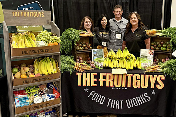 The FruitGuys employees smiling behind a conference booth filled with fresh produce and packaged snacks