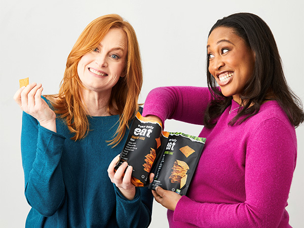 Two smiling women eating packaged snacks