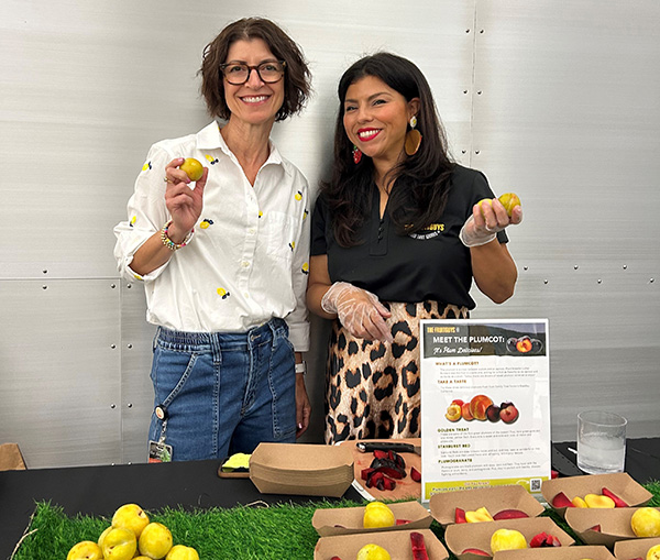 Smiling women holding fresh fruit in front of a table of fruits samples