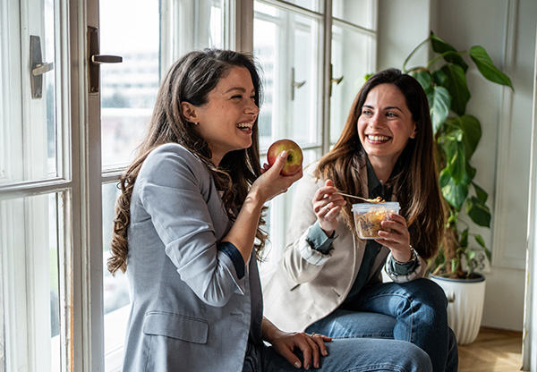 Two smiling businesswomen enjoying a healthy lunch break together, including fruit, sitting by the window in their office
